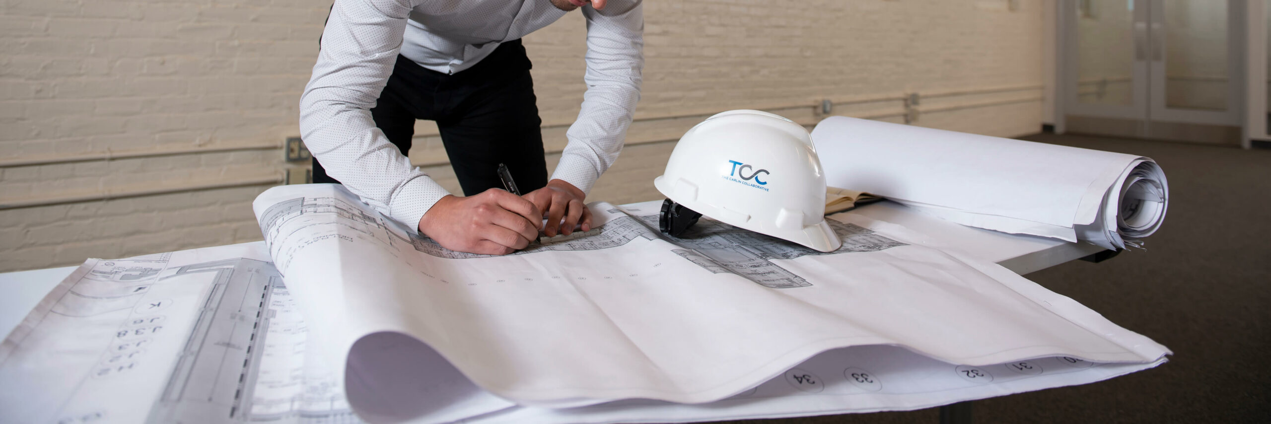 A man in a hard hat and white shirt is focused on working on a blueprint, displaying professionalism and dedication.